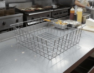Large stainless steel wire mesh basket with side handles on a commercial kitchen prep counter with cooking equipment in the background