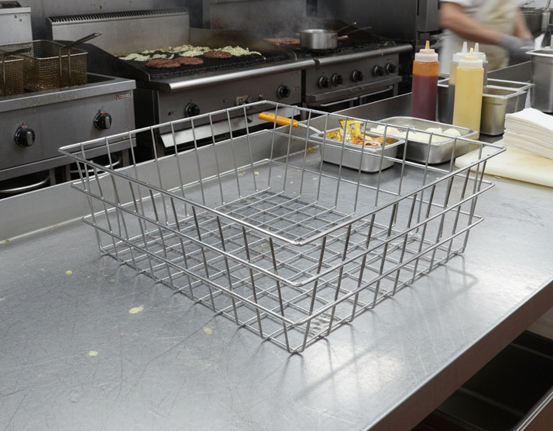Large stainless steel wire mesh basket with side handles on a commercial kitchen prep counter with cooking equipment in the background
